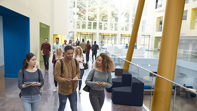 Studierende gehen durch eine Eingangshalle mit bunten Nischen und ockerfarbenen Säulen