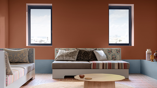 Ein warmes Wohnzimmer mit terrakottafarbenen Wänden und hellblauen Akzenten im unteren Bereich, mit neutralen Möbeln, bunt gemusterten Kissen und einem hellen Couchtisch aus Holz auf einem terrakottafarben gefliesten Boden.